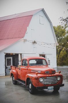 an old red truck is parked in front of a white barn with a red roof