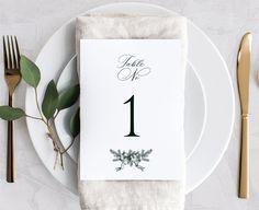 a place setting with white plates and silverware, gold cutlery and greenery