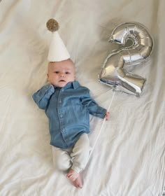 a baby wearing a party hat laying on top of a bed next to the number 2 balloon