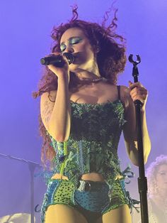 a woman in green and black outfit holding a microphone on stage with other people behind her