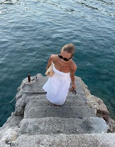 a woman in a white dress sitting on the edge of a cliff next to water