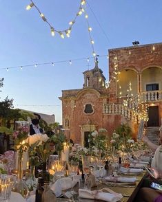 people are sitting at a long table in front of a building with lights strung over it