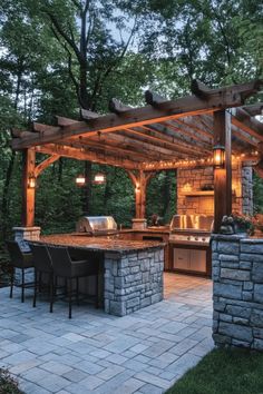 Outdoor kitchen with stone countertops and a pergola, nestled in a wooded area. Outdoor Cooking Area