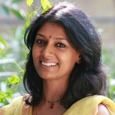 a woman in a yellow sari smiling for the camera with green leaves behind her