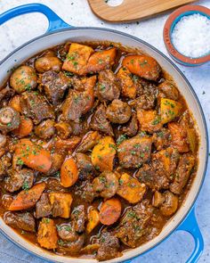 beef stew with carrots and potatoes in a blue pot on a white surface next to a wooden spoon