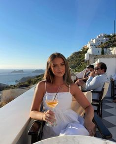 a woman holding a glass of wine sitting on top of a table next to the ocean