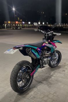 a colorful dirt bike parked on top of a brick road at night with street lights in the background