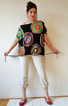 a woman standing in front of a wall with her hands on her hips, wearing white pants and a black top with multicolored circles