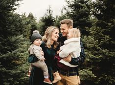 a man and woman holding two small children in their arms while standing next to trees