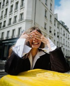 a woman covering her eyes while sitting on top of a yellow car in front of a tall building