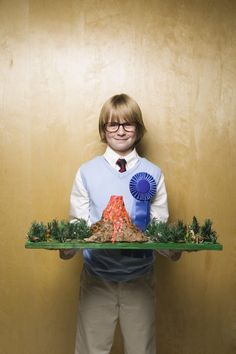 a young boy holding a tray with fake volcanos and trees on it in front of a wall