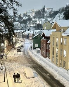 two people walking down a snow covered street in the middle of some buildings and cars