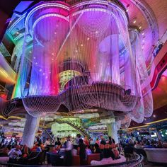 the inside of a casino with people sitting at tables and colorful lights hanging from the ceiling