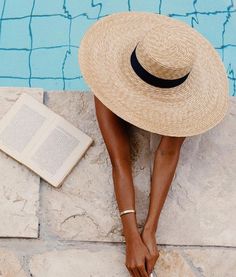 a woman sitting on the edge of a swimming pool wearing a straw hat and reading a book