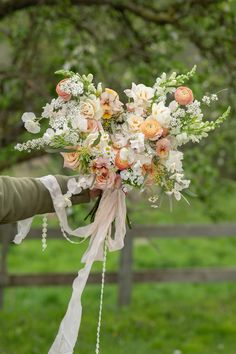 a bridal bouquet with peach and white flowers is held by a woman in front of a fence