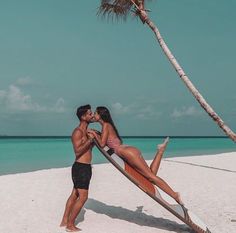 a man and woman kissing on the beach with a surfboard leaning up against a palm tree