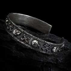 The inspiration for this sterling silver gothic cuff bracelet came from a visit at the Sedlec Ossuary in Czech Republic when my band toured through Europe with Joe Satriani. This small ossuary contains skeleons of betewen 40,000 - 70,000 people artistically arranged into decorations and furnishings. It's truly a stunning macabre location. It features intricate detailing of skulls and bones shaped into a cross encircling the entirety of the bracelet. The frame of the Catacomb Bracelet is designed to resemble the brickwork of the Ossuary's surroundings, adding a touch of realism to this macabre piece. Each skull and femur is expertly crafted to ensure a sense of realism, making the bracelet perfect for those who love to embrace their dark side. I designed this as a matching set to pair with Gothic Silver Adjustable Wristband, Gothic Adjustable Silver Wristband, Adjustable Silver Gothic Wristband, Silver Gothic Adjustable Wristband, Gothic Style Adjustable Silver Wristband, Silver Gothic Skull Bracelets, Silver Punk Wristband As Gift, Silver Punk Style Wristband Gift, Silver Punk Bangle Bracelet