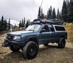 a truck parked on top of a dirt road next to trees and snow covered mountains