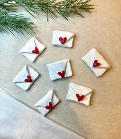 small white envelopes with red hearts are sitting on a table next to a pine branch