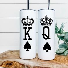 two white tumblers with black letters and crowns are sitting on a wooden table next to a potted plant