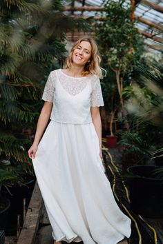 a woman standing in a greenhouse wearing a white dress and smiling with her hands on her hips