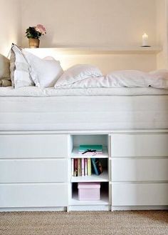 a white bed with bookshelf and drawers underneath it in a room that has carpeted flooring