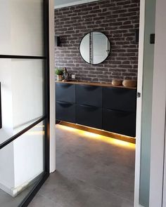 an open door leading to a bathroom with brick wall and black cabinetry, along with a round mirror on the wall