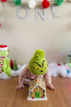 a baby is wearing a green hat while sitting on the floor in front of a gingerbread house