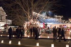 many people are walking around in front of some houses with christmas lights on the trees