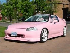 a pink sports car parked in front of a brick driveway with trees and houses behind it