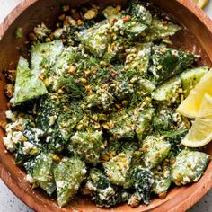 a wooden bowl filled with green vegetables and topped with feta cheese next to a lemon wedge