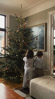 two children standing in front of a christmas tree with lights on it and looking out the window