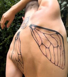 a man with a butterfly wing tattoo on his back and shoulder is standing in front of some bushes