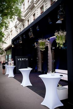 white tables and chairs in front of a building with people walking past them on the sidewalk