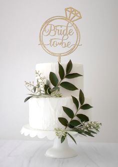 a white wedding cake with greenery and a gold bride to be sign on top