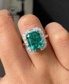 a woman's hand holding an emerald and diamond ring in her left hand, with the center stone surrounded by diamonds