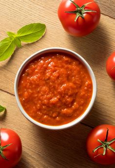 tomato sauce in a white bowl surrounded by tomatoes and green leaves on a wooden table