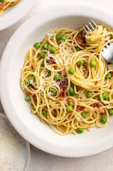 two white bowls filled with pasta and peas on top of a marble table next to silver utensils