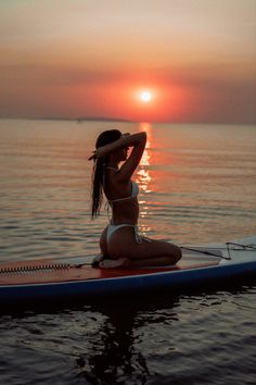 a woman sitting on top of a surfboard in the ocean at sunset with her arms behind her head