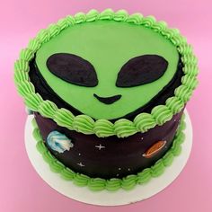 a green alien cake with black eyes and stars on it's face is sitting on a pink surface