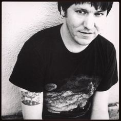 black and white photograph of a young man with tattoos on his arms sitting against a wall