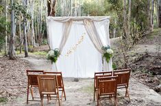 two wooden chairs are set up in front of a white drape with flowers and greenery