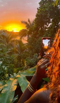 a woman taking a photo with her cell phone in front of the sun setting over some trees