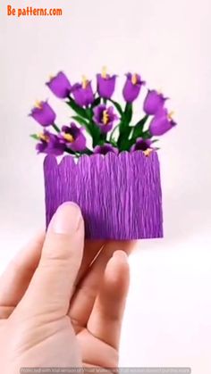 a hand holding a small purple flower pot with flowers in it's bottom half