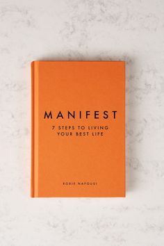 an orange book sitting on top of a white counter next to a marble wall with the title'manfest 7 steps to living your best life '