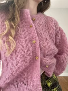 a woman wearing a pink cardigan and plaid pants standing in front of a white wall