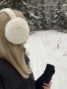 a woman standing in the snow with ear muffs on her head looking at trees