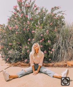 a woman sitting on the ground in front of a bush with pink flowers behind her