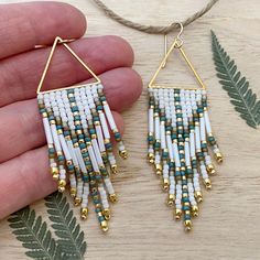 a pair of white and gold beaded earrings on a wooden table with fern leaves