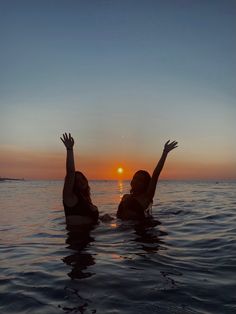 two women in the water with their arms up at sunset or sunrise, one raising her hand to the sky